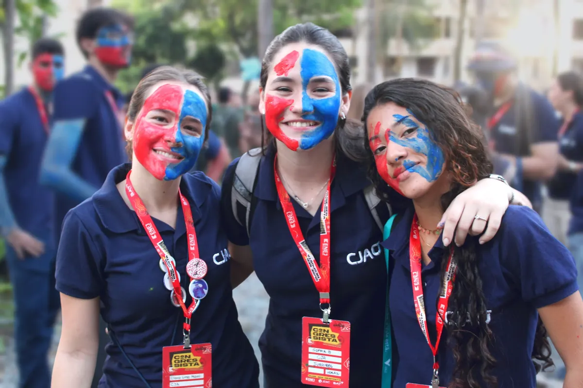 Três membros da CJA abraçados, com o rosto pintado metade de vermelho e metade de azul.
