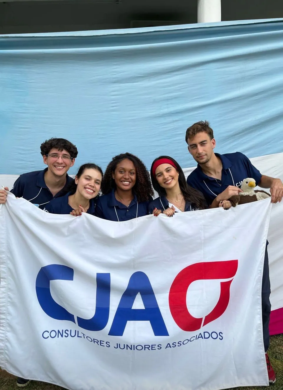 Cinco integrantes da CJA de polo azul-marinho segurando a bandeira da CJA, em frente à bandeira do Espírito Santo.