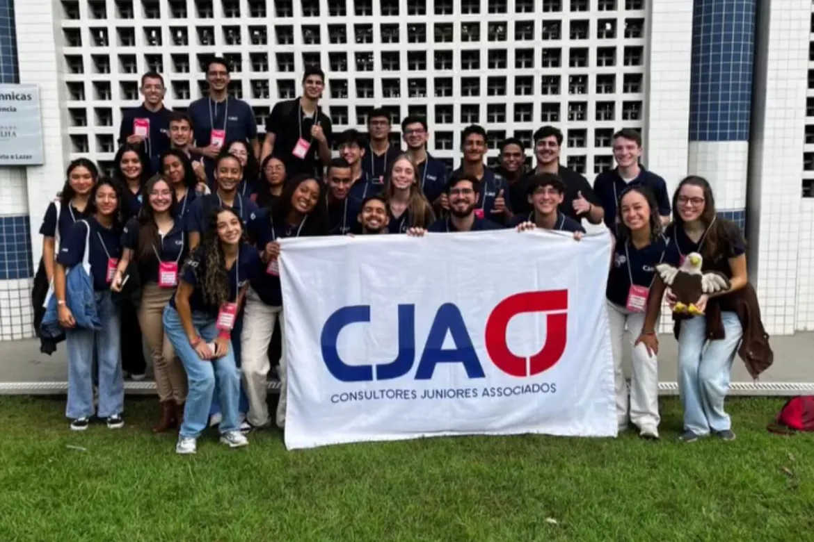 O time da CJA, de polo azul-marinho, reunido com a bandeira da CJA em frente a um prédio da UFES.