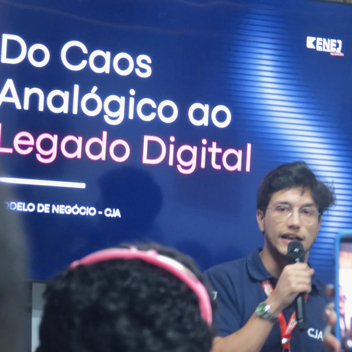 Um membro da CJA, de polo azul-marinho, fala ao microfone diante do slide Do Caos Analógico ao Legado Digital