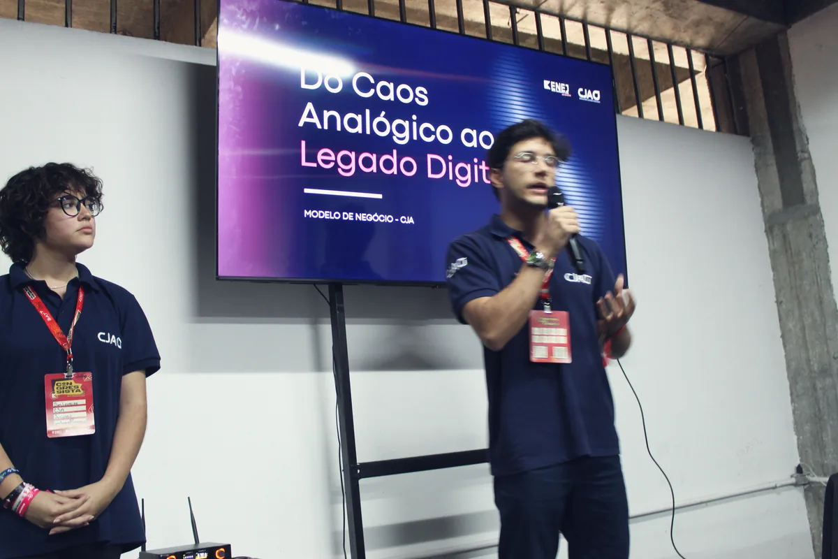 Dois membros da CJA, de polo azul-marinho, diante de um slide; um deles apresenta ao microfone